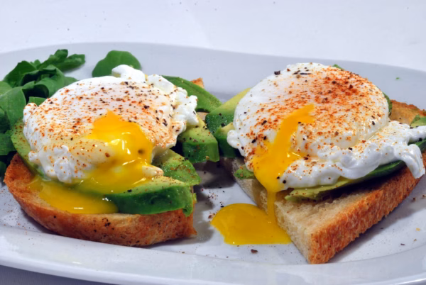 Two slices of toast topped with avocado and poached eggs, sprinkled with seasoning. One egg has a runny yolk spilling onto the toast. Some leafy greens are on the side of the white plate.