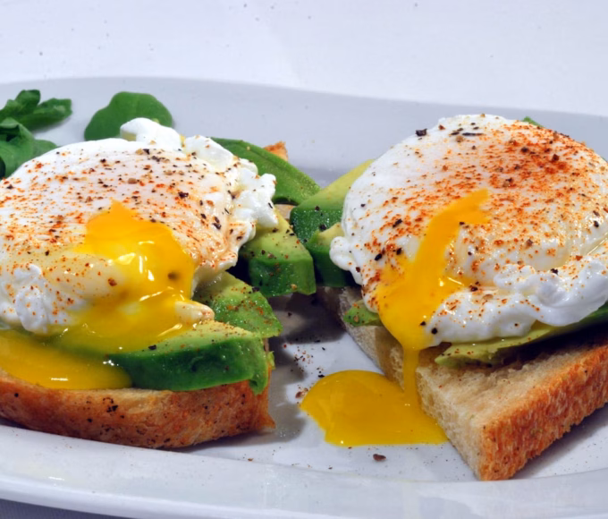 Two slices of toast topped with avocado and poached eggs, sprinkled with seasoning. One egg has a runny yolk spilling onto the toast. Some leafy greens are on the side of the white plate.