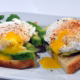 Two slices of toast topped with avocado and poached eggs, sprinkled with seasoning. One egg has a runny yolk spilling onto the toast. Some leafy greens are on the side of the white plate.