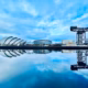 A cityscape featuring modern buildings, a crane, and a domed structure, all reflected clearly in a calm body of water under a bright blue sky with scattered clouds.