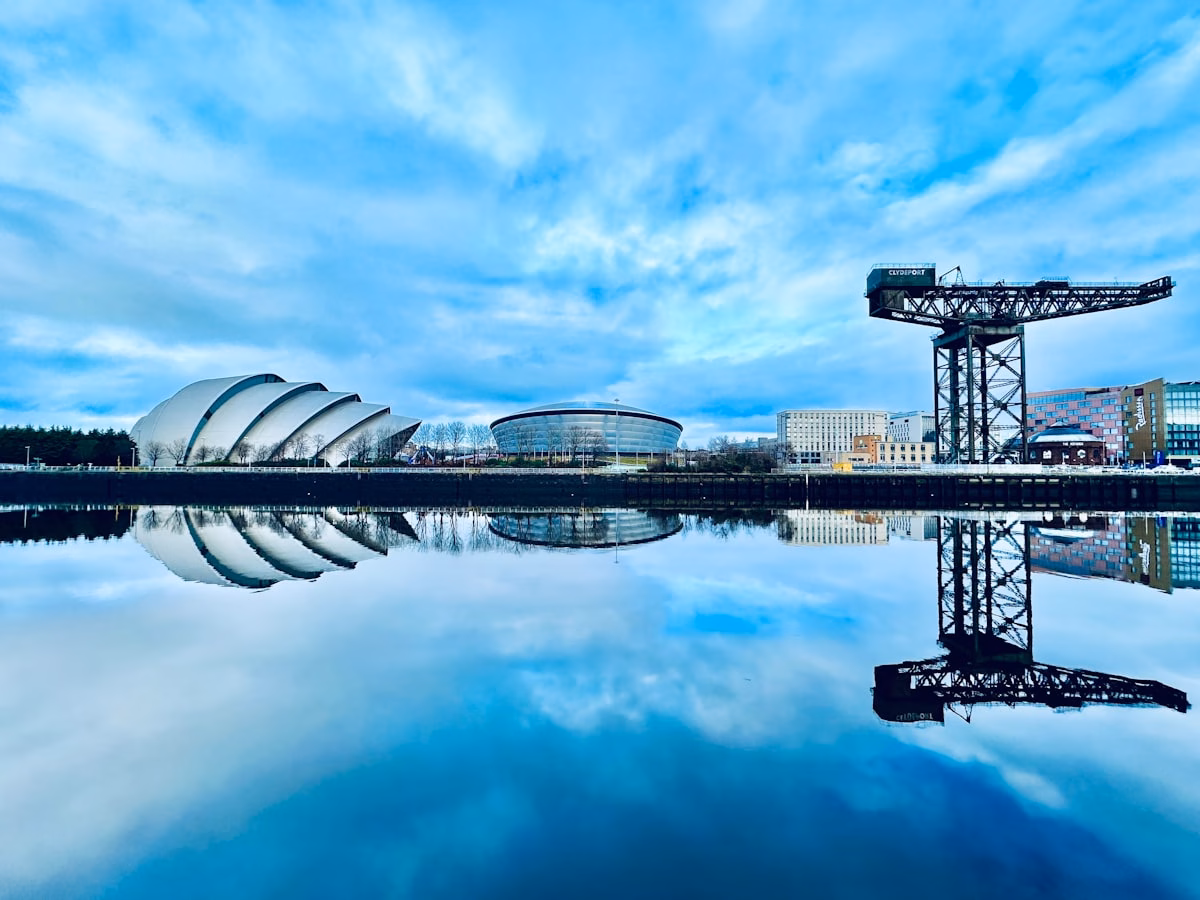 A cityscape featuring modern buildings, a crane, and a domed structure, all reflected clearly in a calm body of water under a bright blue sky with scattered clouds.