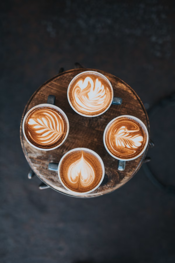 Four mugs of coffee with different intricate latte art designs are arranged in a circle on a rustic wooden table, viewed from above.