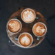 Four mugs of coffee with different intricate latte art designs are arranged in a circle on a rustic wooden table, viewed from above.