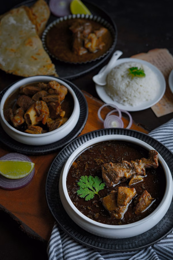 Indian meal with bowls of mutton curry and goat stew, served with rice, flatbread, sliced red onions, a lemon wedge, and garnished with fresh coriander on a dark rustic background.