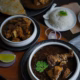 Indian meal with bowls of mutton curry and goat stew, served with rice, flatbread, sliced red onions, a lemon wedge, and garnished with fresh coriander on a dark rustic background.