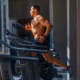 A muscular man runs on a treadmill in a gym, wearing black shorts and trainers. Sunlight streams through large windows, illuminating the workout area and highlighting the man's physique.