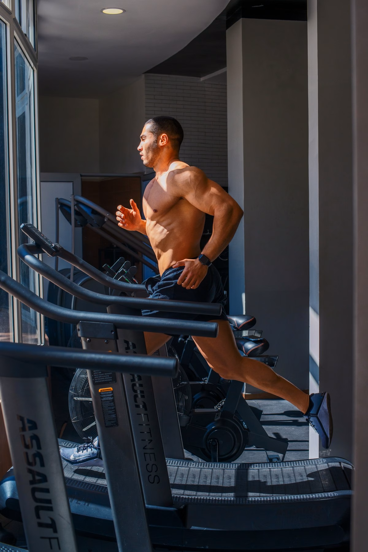 A muscular man runs on a treadmill in a gym, wearing black shorts and trainers. Sunlight streams through large windows, illuminating the workout area and highlighting the man's physique.