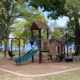 Children play on a playground with slides, swings, and climbing frames surrounded by trees. In the background, adults sit at picnic tables near a lake under a clear blue sky.