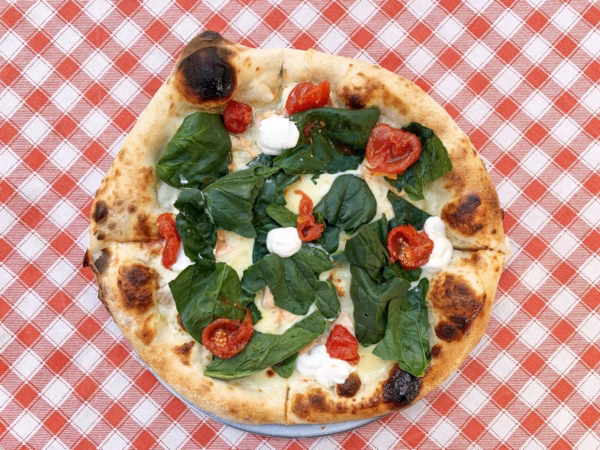 A pizza with fresh spinach, roasted red peppers, melted cheese, and dollops of white sauce sits on a white plate atop a red-and-white chequered tablecloth.