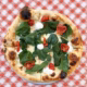 A pizza with fresh spinach, roasted red peppers, melted cheese, and dollops of white sauce sits on a white plate atop a red-and-white chequered tablecloth.