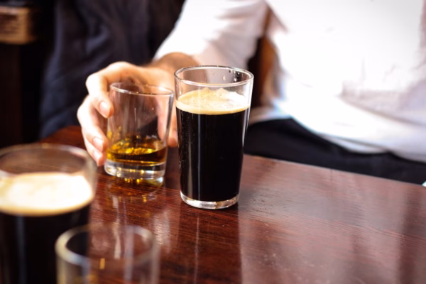 A person wearing a white shirt holds a glass of whisky next to a pint of dark beer on a wooden table, with another pint partially visible in the foreground.