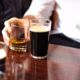 A person wearing a white shirt holds a glass of whisky next to a pint of dark beer on a wooden table, with another pint partially visible in the foreground.