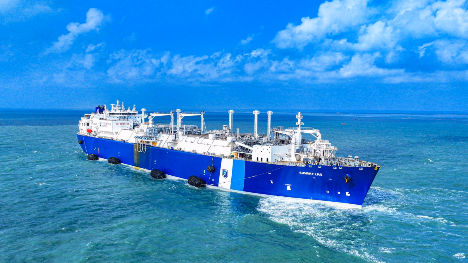 A large blue and white Summit Group liquefied natural gas (LNG) tanker sails on calm, turquoise waters under a bright sky, equipped with advanced processing structures and pipes to deliver locked-in fuel at market price.